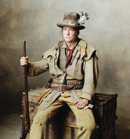 Weigert, dressed as Calamity Jane, sits on a wooden trunk, holding a long-barrel gun. She wears buckskin pants, an gun belt with a pistol in the holster and a row of bullets, a long-sleeve shirt and a hat with a feather stuck in its brim.